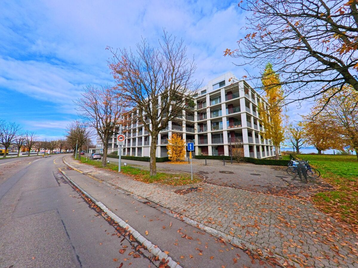 Entlang einer baumbestandenen Strasse in Arbon (9320, Kanton Thurgau) steht eine moderne Wohnueberbauung neben Gruenflaechen mit herbstlich gefaerbten Baeumen.