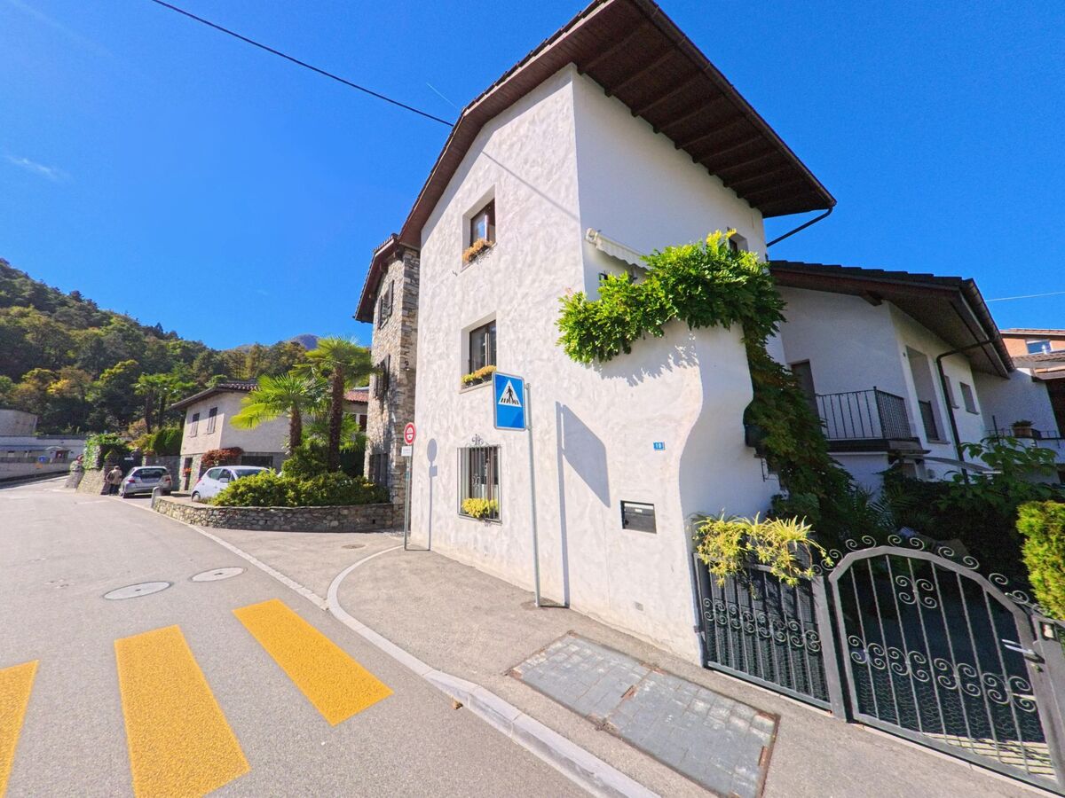 Ein weiss verputztes Haus mit gruenen Pflanzen steht in Arcegno (6616, Kanton Tessin) an einer sonnigen Strasse mit Fussgaengerstreifen.