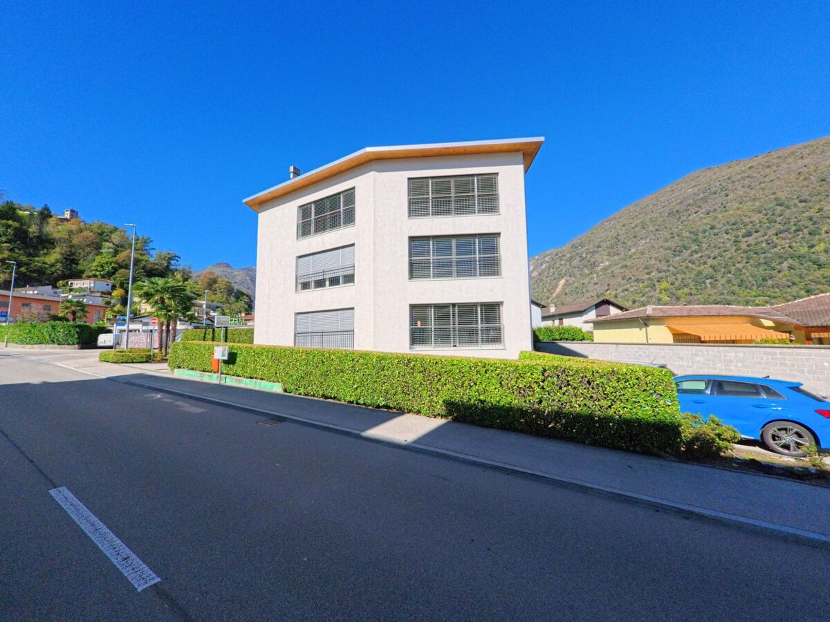 Ein modernes Wohnhaus mit grossen Fenstern steht in Arcegno (6616, Kanton Tessin) vor einer Bergkulisse und gruener Hecke.