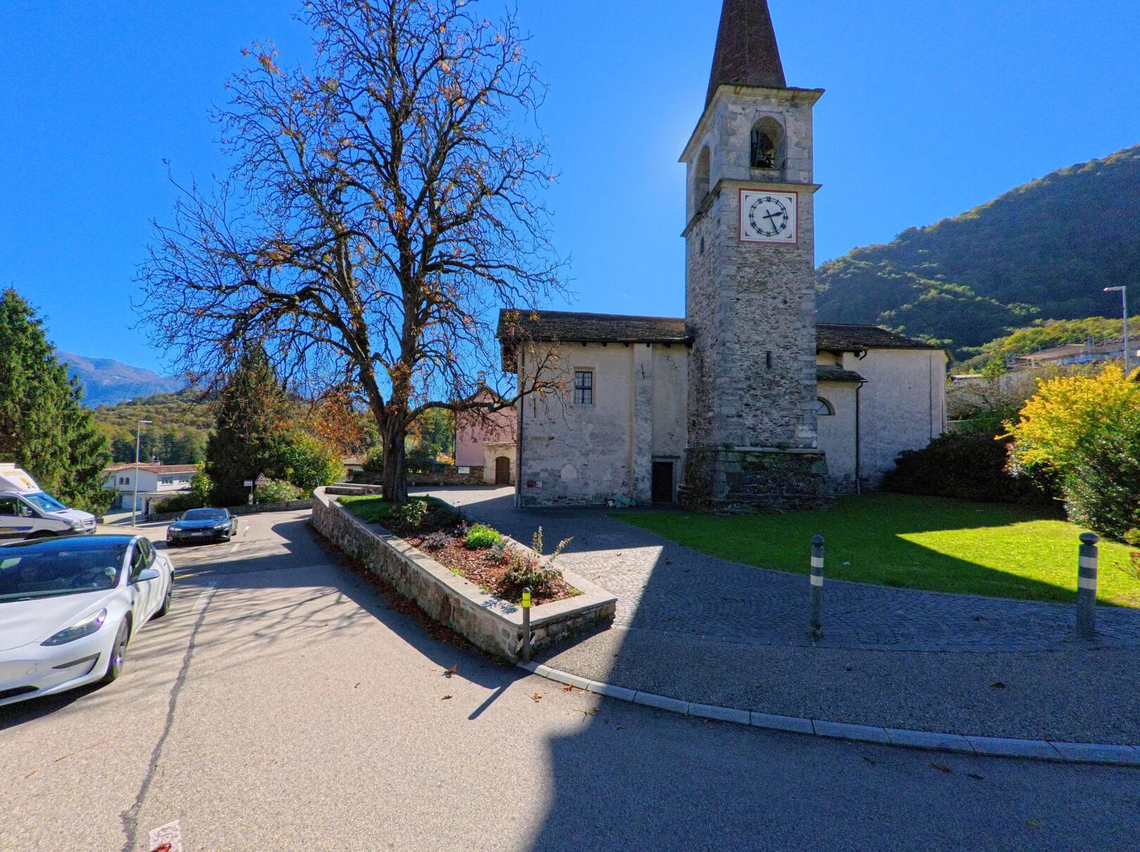 Die steinerne Kirche mit Uhrturm in Arcegno (6618, Kanton Tessin) steht auf einer kleinen Anhöhe, umgeben von Strasse und Bäumen.