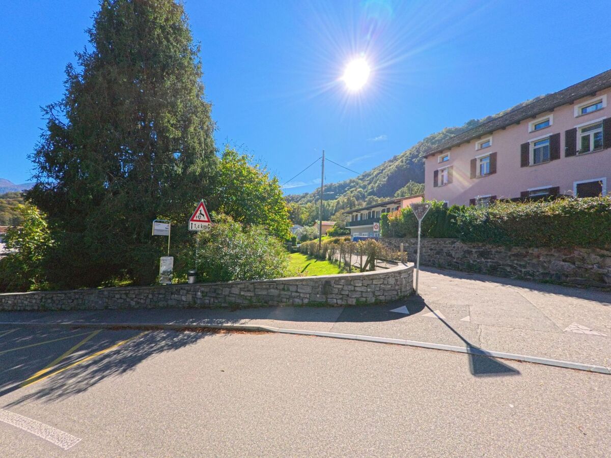 Eine Kreuzung in Arcegno (6618, Kanton Tessin) mit rosa Wohnhaus und grossen Baeumen liegt sonnig am Fusse bewaldeter Huegel.
