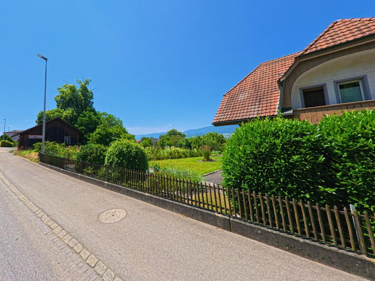 In Arch (3296, Kanton Bern) verläuft eine ruhige Anliegerstrasse mit gepflegten Vorgärten und Blick auf die Jurakette am Horizont.