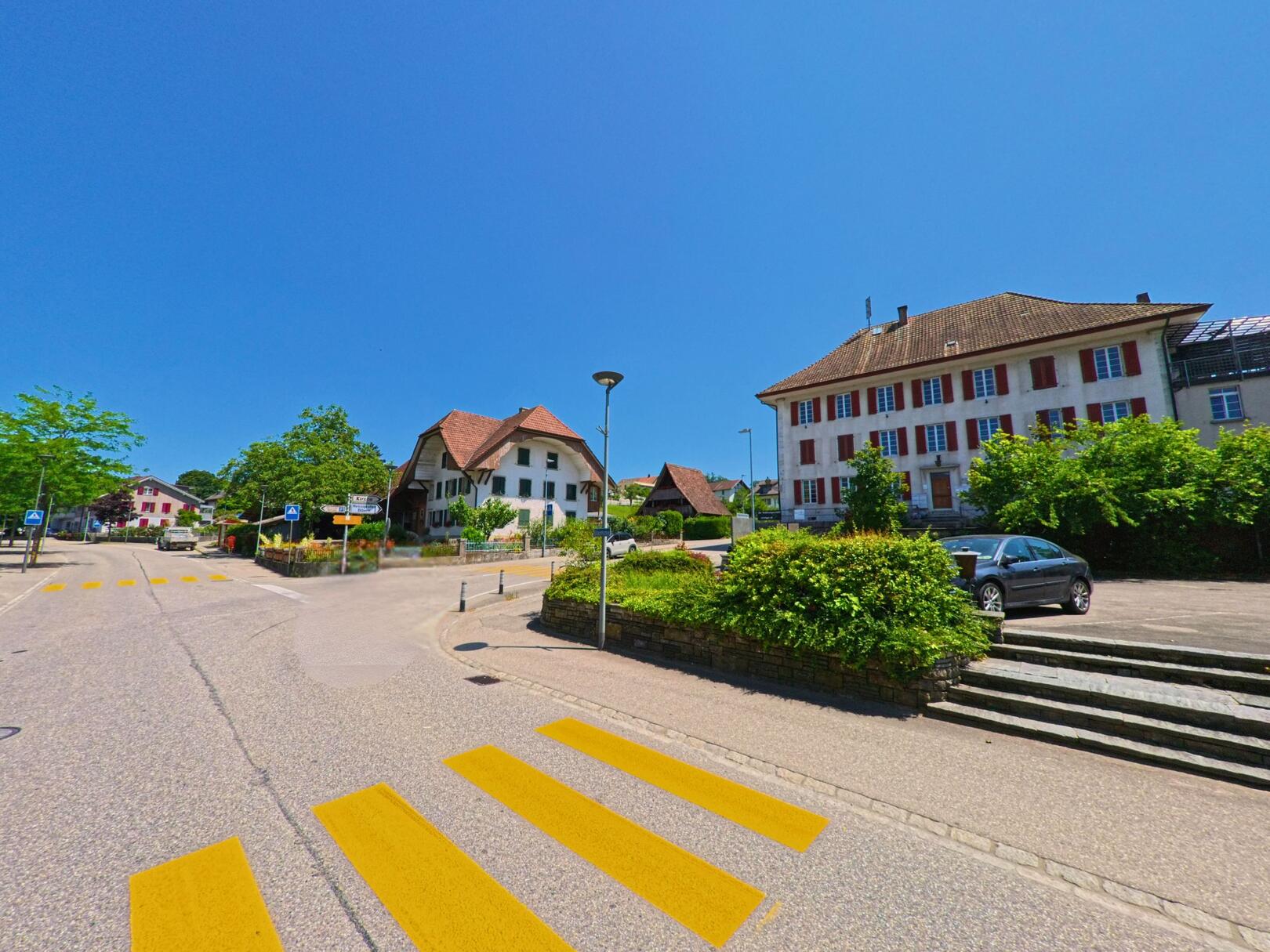 In Arch (PLZ 3296, Kanton Bern) prägt ein historisches Gebäude mit roten Fensterläden das Dorfbild an einer verkehrsberuhigten Kreuzung mit Zebrastreifen.
