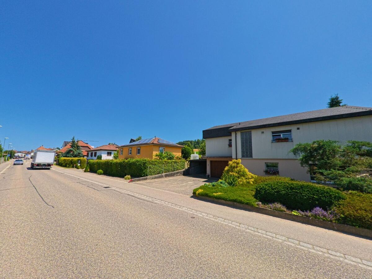 Entlang einer Durchgangsstrasse in Arch (PLZ 3296, Kanton Bern) stehen Einfamilienhäuser mit gepflegten Gärten, während Autos die sonnige Strecke befahren.