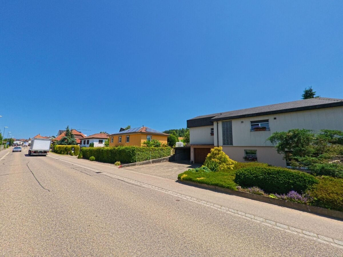 Entlang einer Durchgangsstrasse in Arch (PLZ 3296, Kanton Bern) stehen Einfamilienhäuser mit gepflegten Gärten, während Autos die sonnige Strecke befahren.