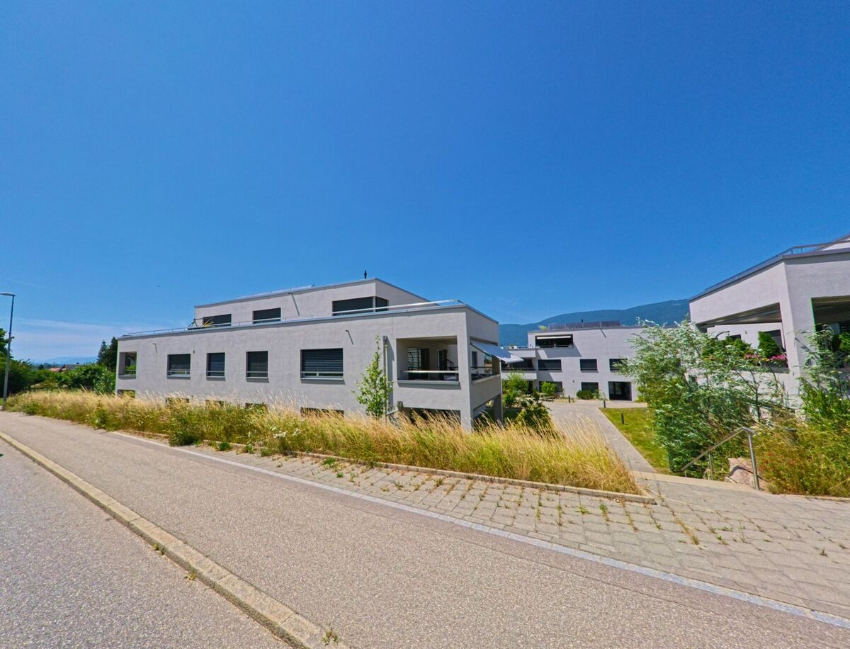 In einem neueren Wohnquartier von Arch (PLZ 3296, Kanton Bern) stehen moderne Mehrfamilienhäuser mit Flachdach entlang eines sanft ansteigenden Wegs.