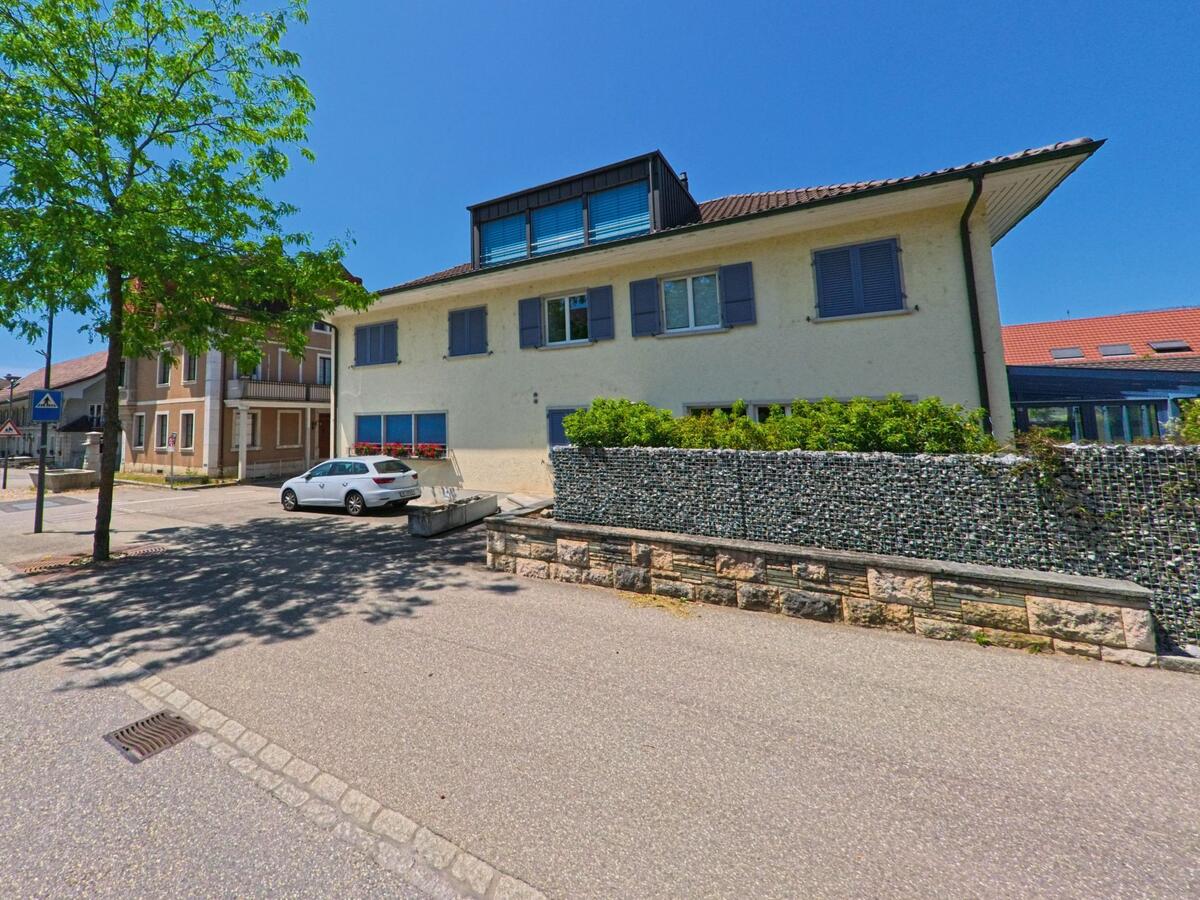 In Arch (PLZ 3296, Kanton Bern) säumen Wohnhäuser mit Fensterläden eine ruhige Seitenstrasse, vor der ein Auto unter einem schattenspendenden Baum parkt.