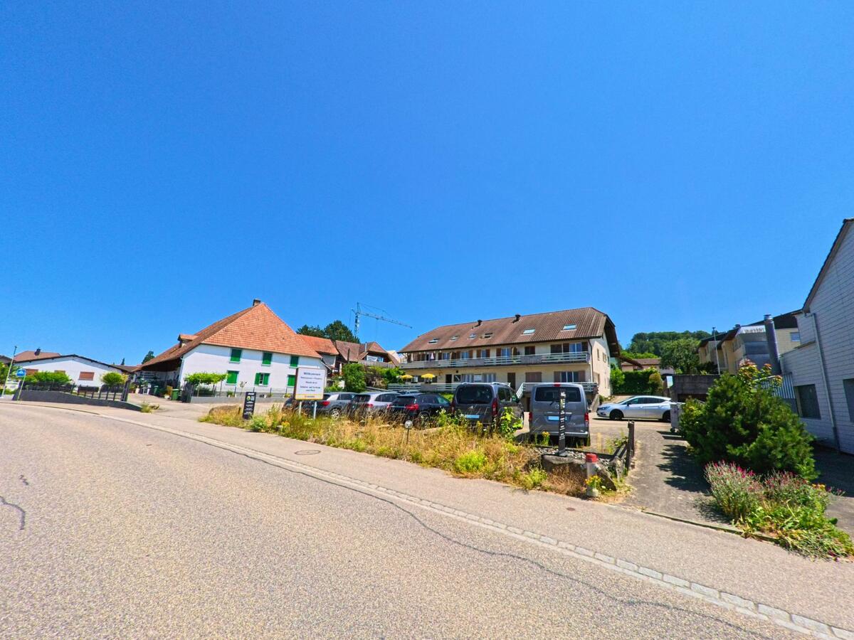 In Arch (3296, Kanton Bern) reihen sich entlang einer Hauptstrasse Wohnhäuser mit Parkplätzen und begrünten Vorgärten vor bewaldetem Hügelland.