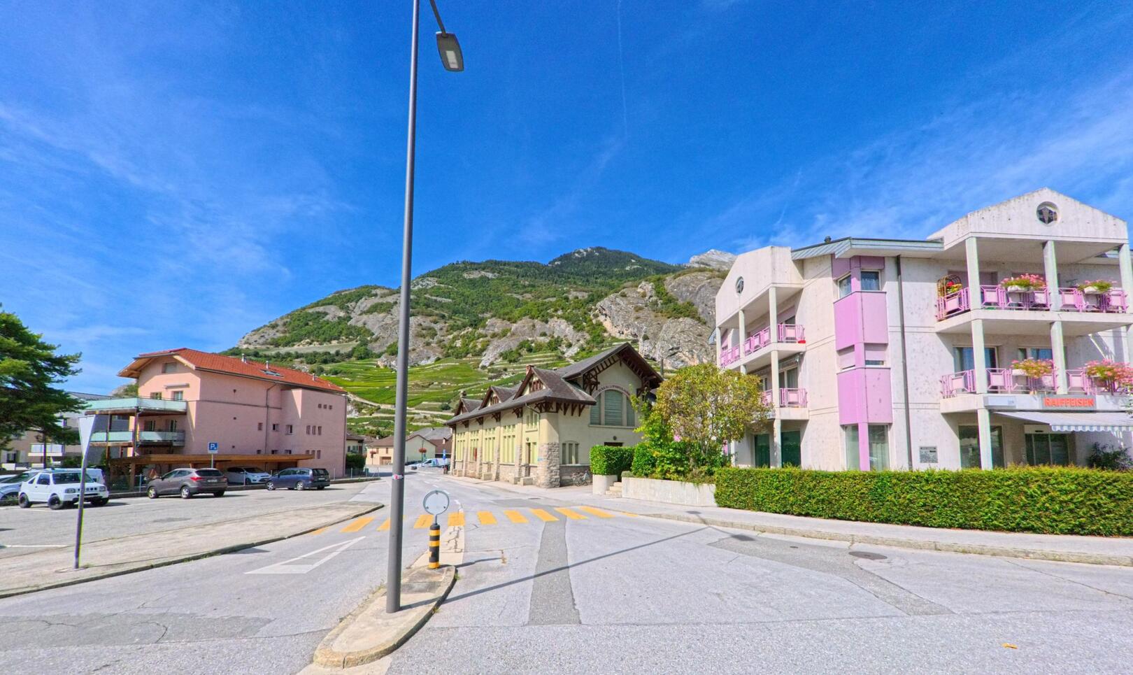 In Ardon (1957, Kanton Wallis) stehen ein modernes Wohnhaus mit rosa Balkonen und ein historisches Gebaeude vor den Weinbergen am Hang.