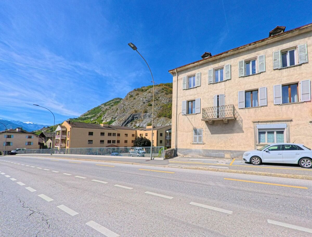 Ein Wohnhaus mit Balkonen und Fensterlaeden steht in Ardon (1957, Kanton Wallis) direkt an der Hauptstrasse vor dem felsigen Hang.
