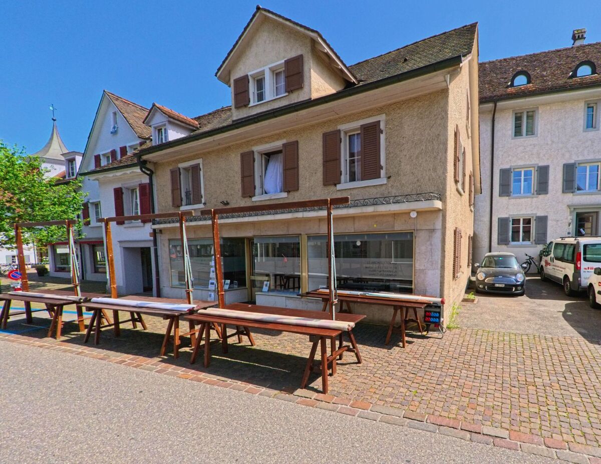 In Arlesheim (4144, Kanton Basel-Landschaft) stehen entlang einer gepflasterten Gasse mehrere historische Wohn- und Geschäftshäuser mit Fensterläden und Verkaufsständen.