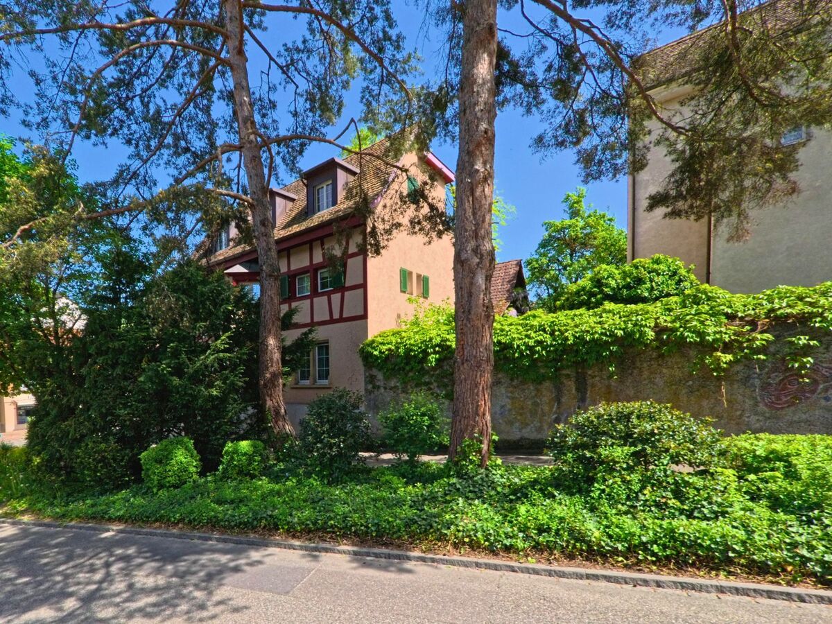 Hinter hohen Bäumen und dichter Vegetation steht in Arlesheim (4144, Kanton Basel-Landschaft) ein Fachwerkhaus mit roten Balken und grünen Fensterläden.