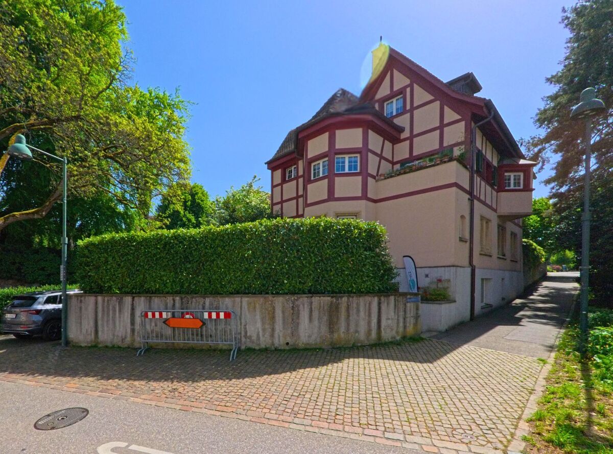 In Arlesheim (4144, Kanton Basel-Landschaft) steht ein Haus im Fachwerkstil mit rot-weissen Balken an einer Kopfsteinpflasterstrasse, umgeben von dichter Hecke und altem Baumbestand.