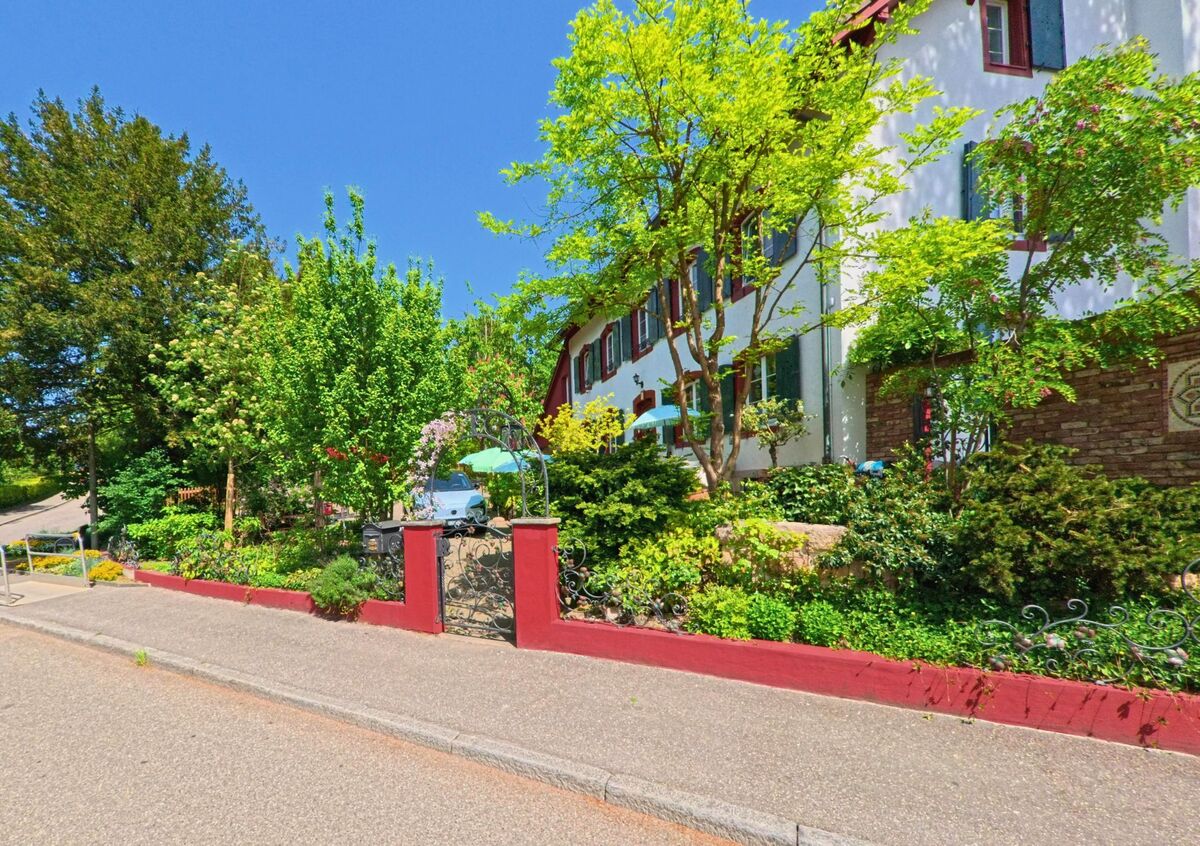 In Arlesheim (4144, Kanton Basel-Landschaft) liegt ein weiss gestrichenes Haus mit roten Fensterläden hinter einem reich bepflanzten Garten und einem verzierten Eisentor.