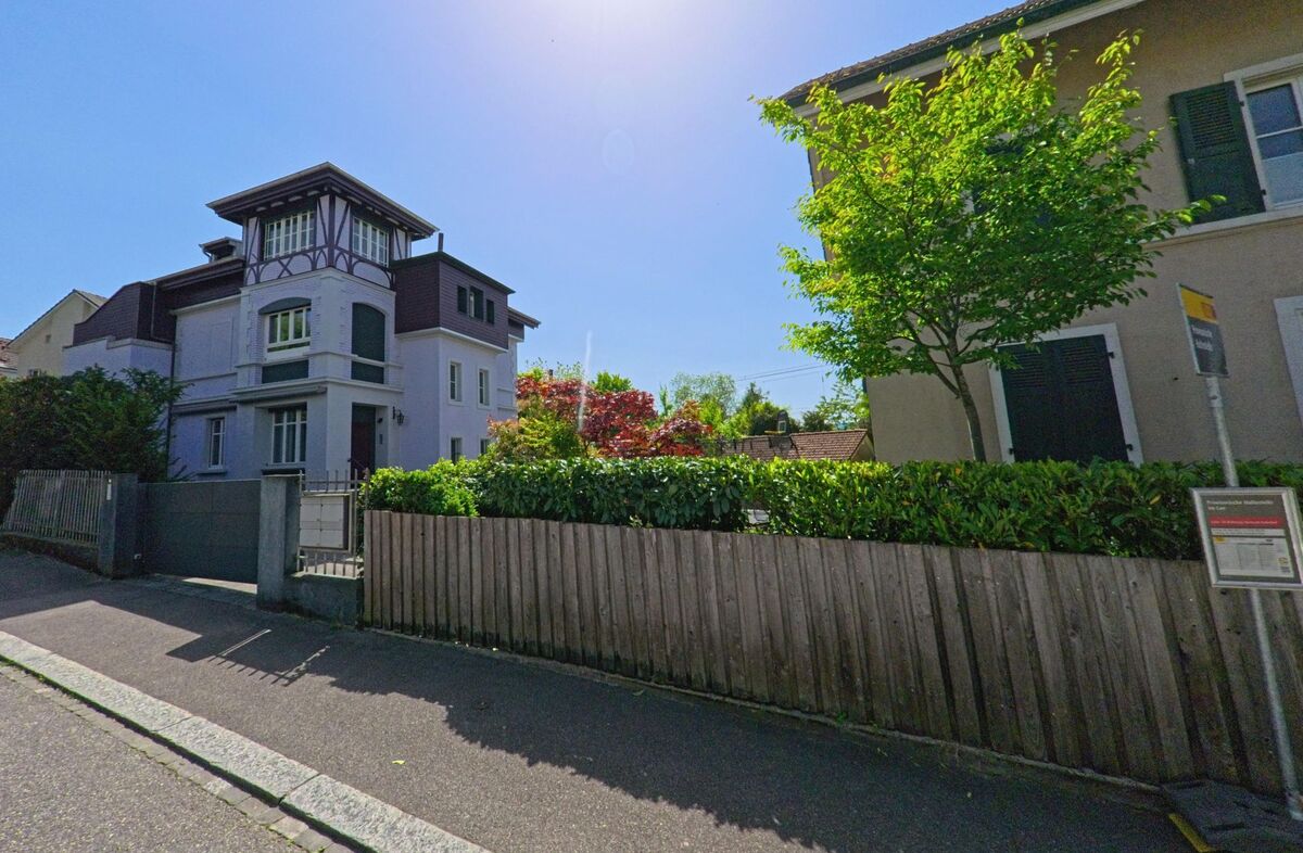 In einem ruhigen Quartier von Arlesheim (4144, Kanton Basel-Landschaft) steht ein mehrstöckiges Wohnhaus mit dekorativen Fassadenelementen hinter einem Holzzaun.