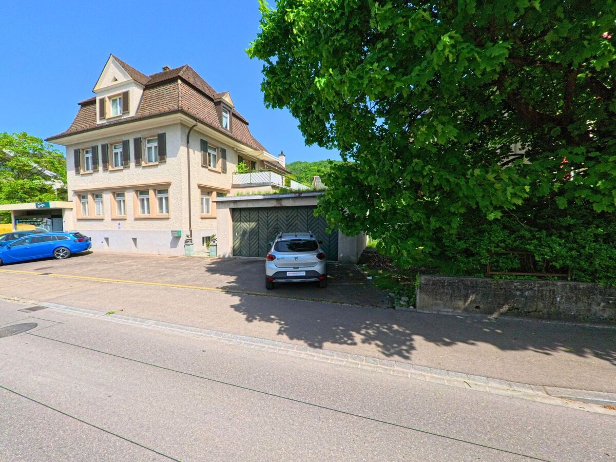 In Arlesheim (4144, Kanton Basel-Landschaft) steht ein gepflegtes Wohnhaus mit markantem Dachfirst neben einer Garage und schattenspendenden Bäumen.