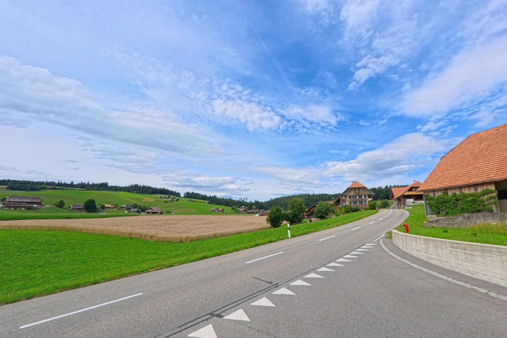 Eine sanft geschwungene Landstrasse führt durch Arni (3508, Kanton Bern) vorbei an Feldern und Bauernhäusern inmitten einer weitläufigen Hügellandschaft.