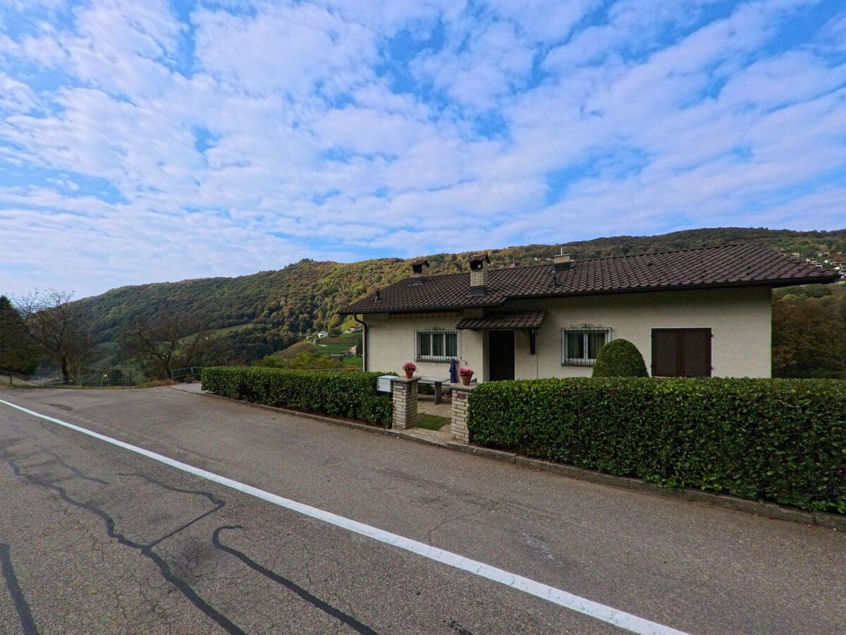 Ein Einfamilienhaus in Arogno (6822, Kanton Tessin) steht ruhig an einer Strasse mit Blick auf bewaldete Hügel und das darunterliegende Tal.
