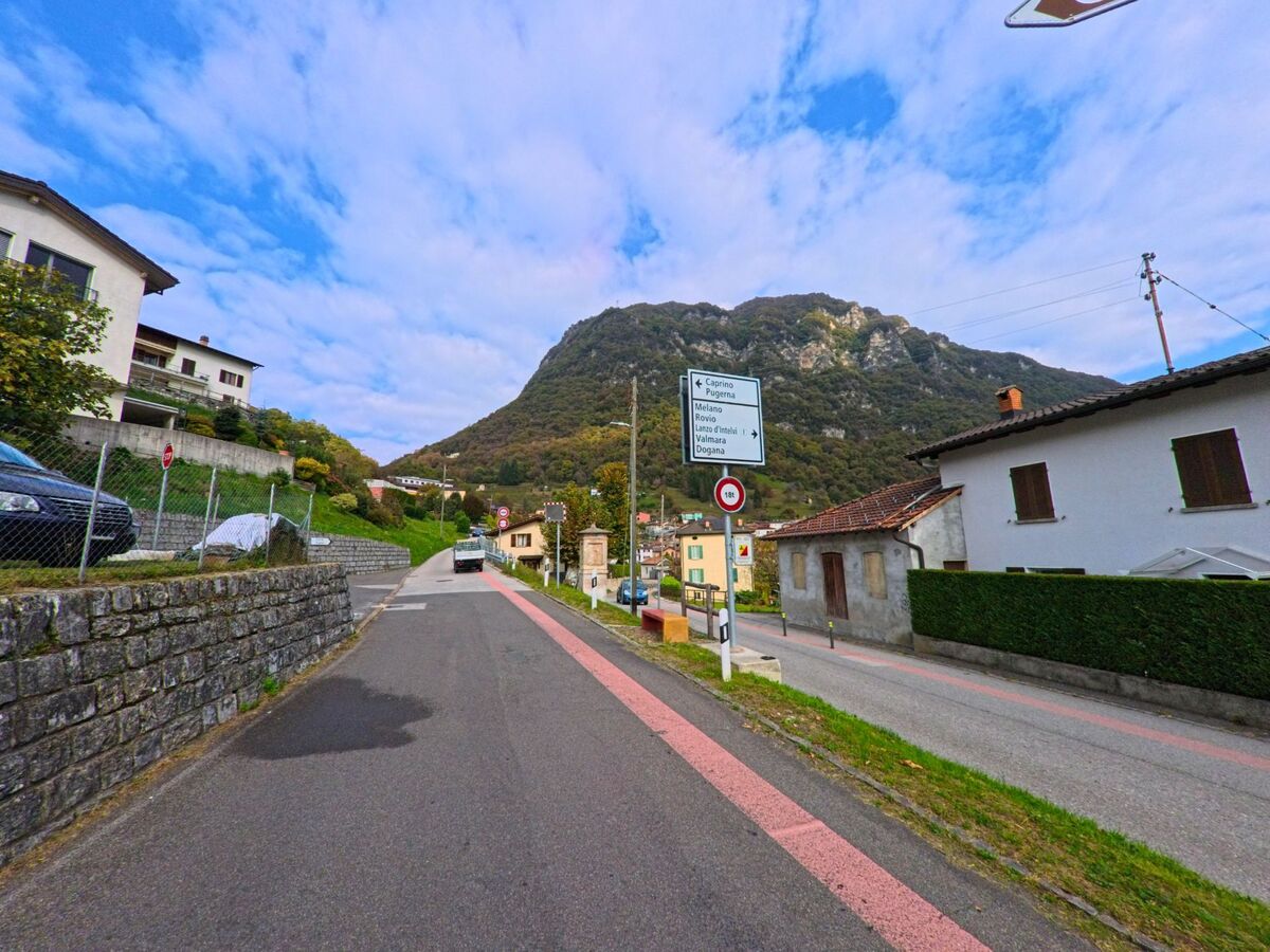 Eine Strasse in Arogno (6822, Kanton Tessin) führt an Wohnhäusern vorbei in Richtung der bewaldeten Hügel, während Wegweiser die Route nach Melano und Caprino anzeigen.