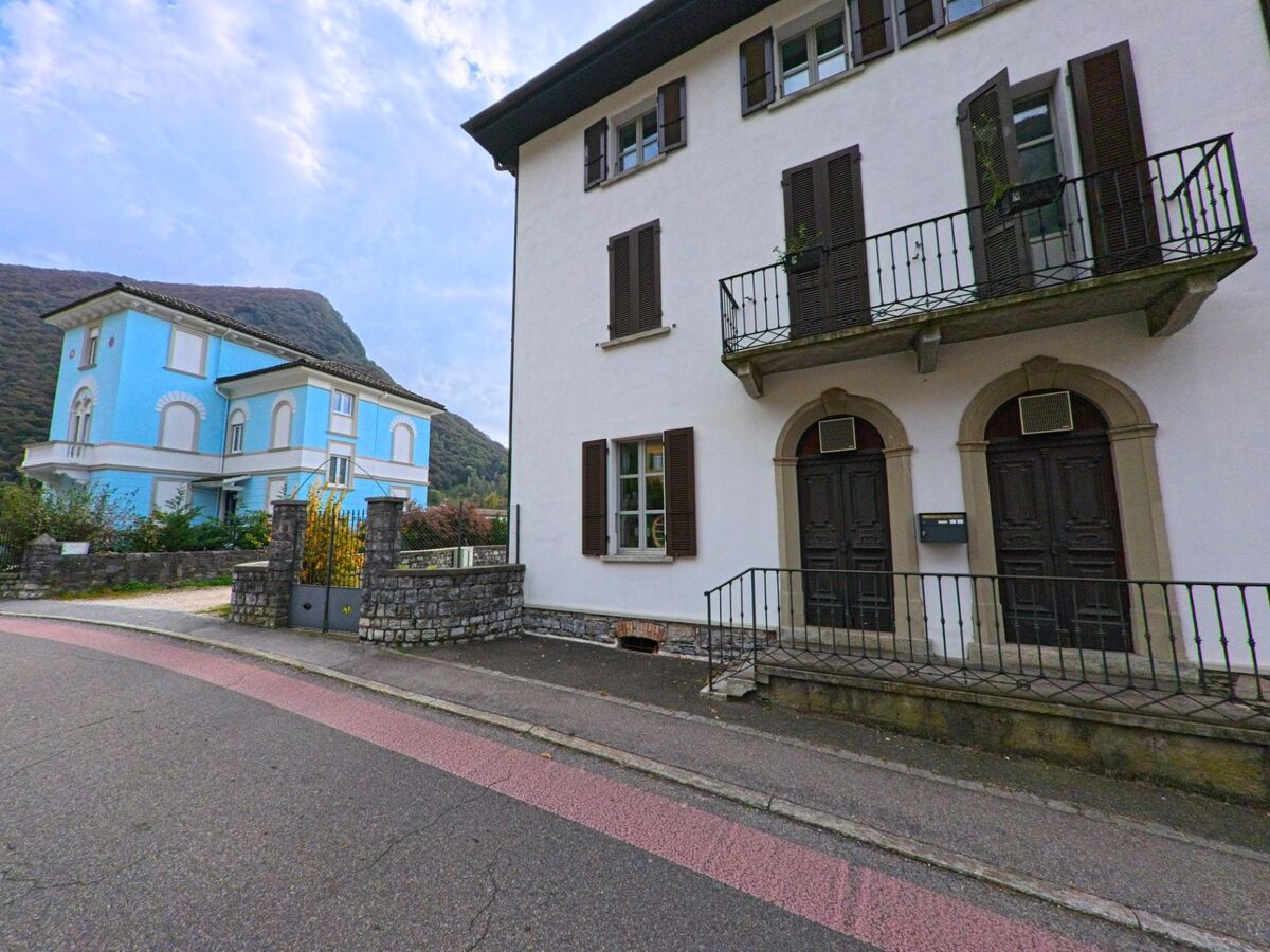 Zwei Wohnhäuser in Arogno (6822, Kanton Tessin), eines in hellblauer Farbe und eines mit dunklen Fensterläden, liegen an einer leicht ansteigenden Strasse am Fuss des Berges.