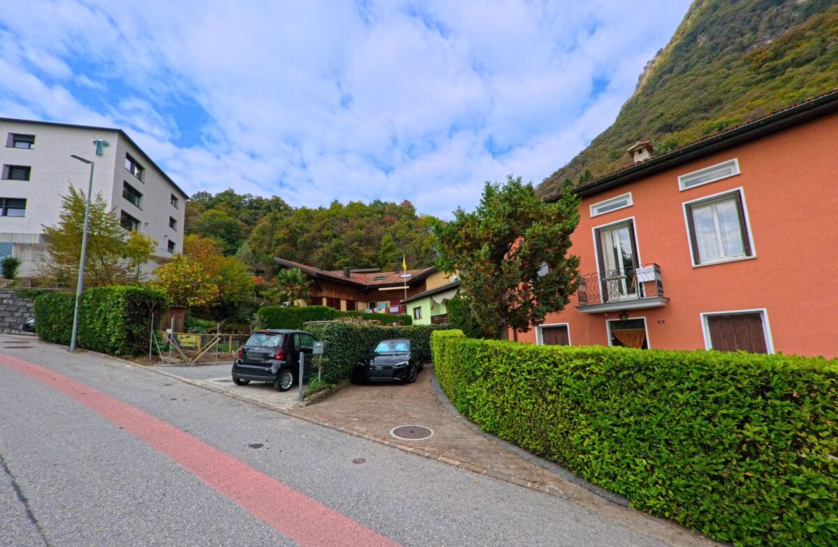 Entlang einer ruhigen Strasse in Arogno (6822, Kanton Tessin) stehen gepflegte Wohnhäuser mit bunten Fassaden und grünen Hecken am Rand eines bewaldeten Hanges.