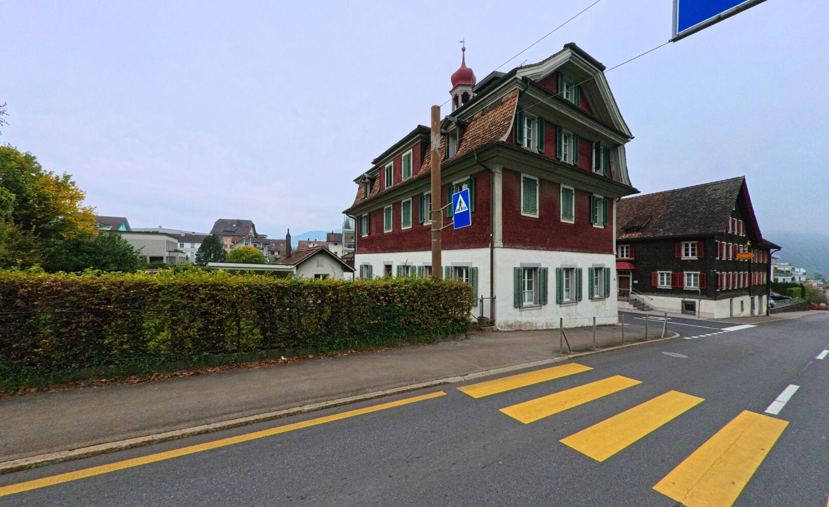 Vor traditionellen Holzh aeusern in Arth (6415, Kanton Schwyz) verlaeuft eine ruhige Strasse mit gelbem Fussgaengerstreifen und Blick auf den leicht bewoelkten Himmel.
