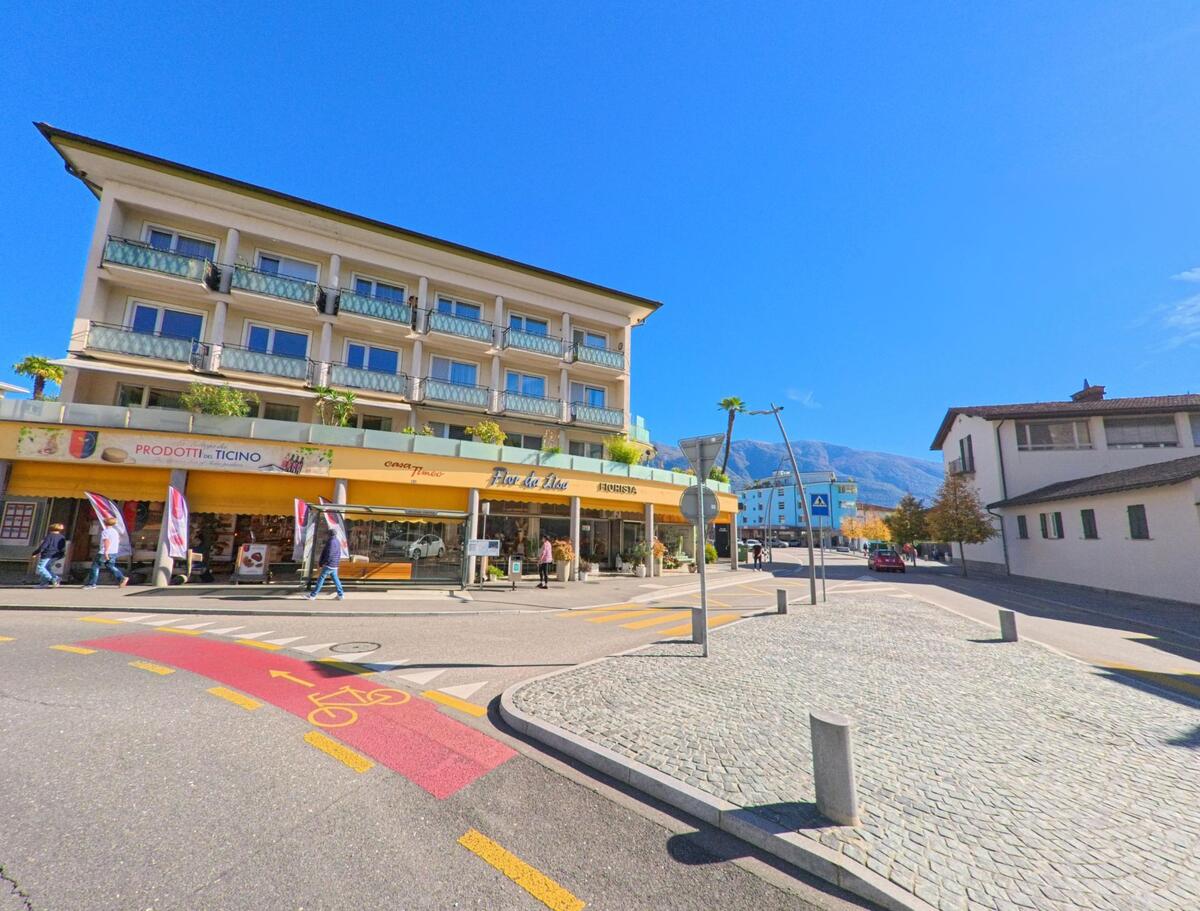 In Ascona (6612, Kanton Tessin) steht an einer sonnigen Kreuzung ein gelbes Gebäude mit Läden und Balkonen vor dem Panorama der Alpen.