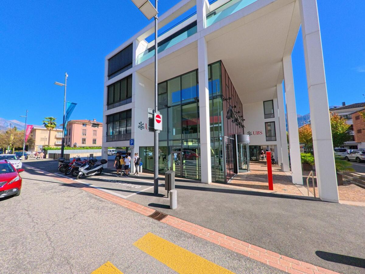 In Ascona (6612, Kanton Tessin) steht ein modernes Bankgebäude mit Glasfassade und hohen Säulen an einer sonnigen Strasse mit Passanten.