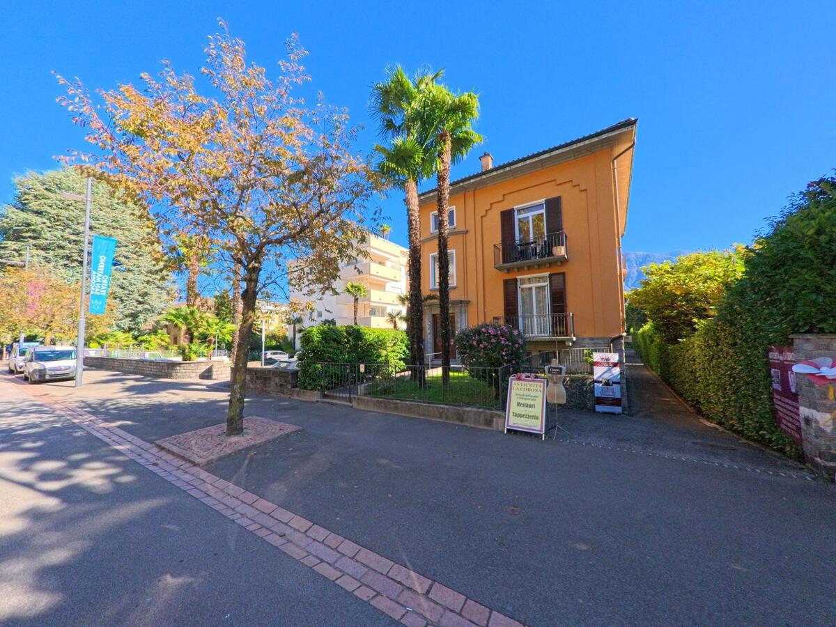 Ein orangefarbenes Wohnhaus mit Palmen und blühenden Sträuchern steht in Ascona (6612, Kanton Tessin) an einer sonnigen, ruhigen Seitenstrasse.