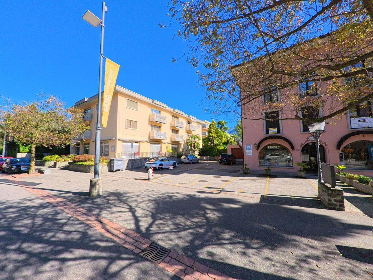 In Ascona (6612, Kanton Tessin) grenzen ein rosafarbenes und ein helles Wohnhaus an einen kleinen Parkplatz mit Schatten spendenden Bäumen.