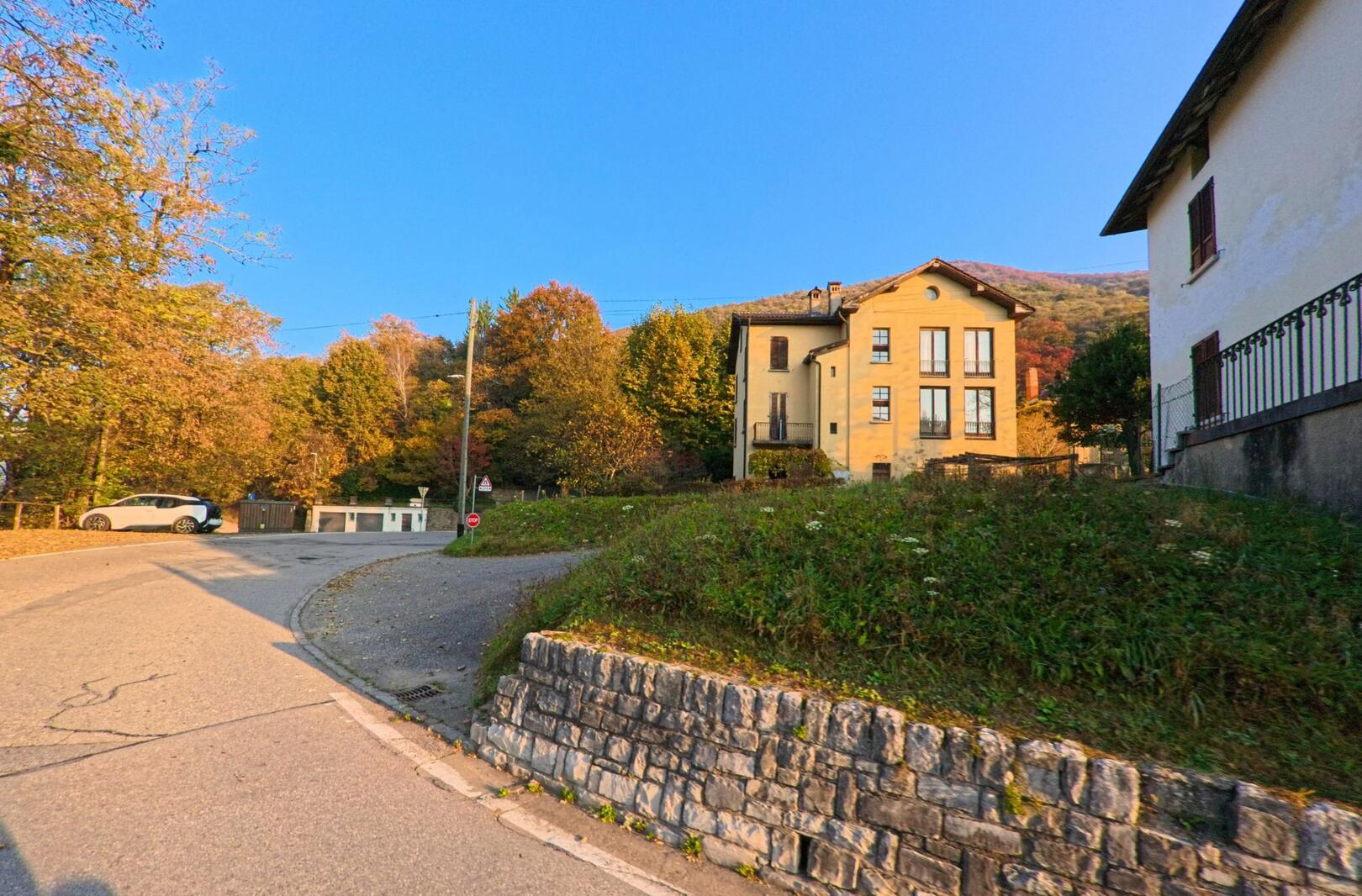 An einer ruhigen Strassenkurve in Astano (6999, Kanton Tessin) stehen Wohnhäuser vor herbstlich gefärbten Bäumen am Hang.