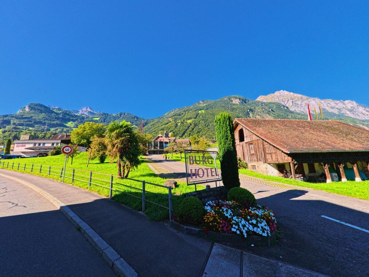 Vor dem Burg Hotel in Attinghausen (6468, Kanton Uri) steht ein Blumenschmuck mit bunten Blumen, daneben ein Holzstadel und die Berglandschaft im Hintergrund.