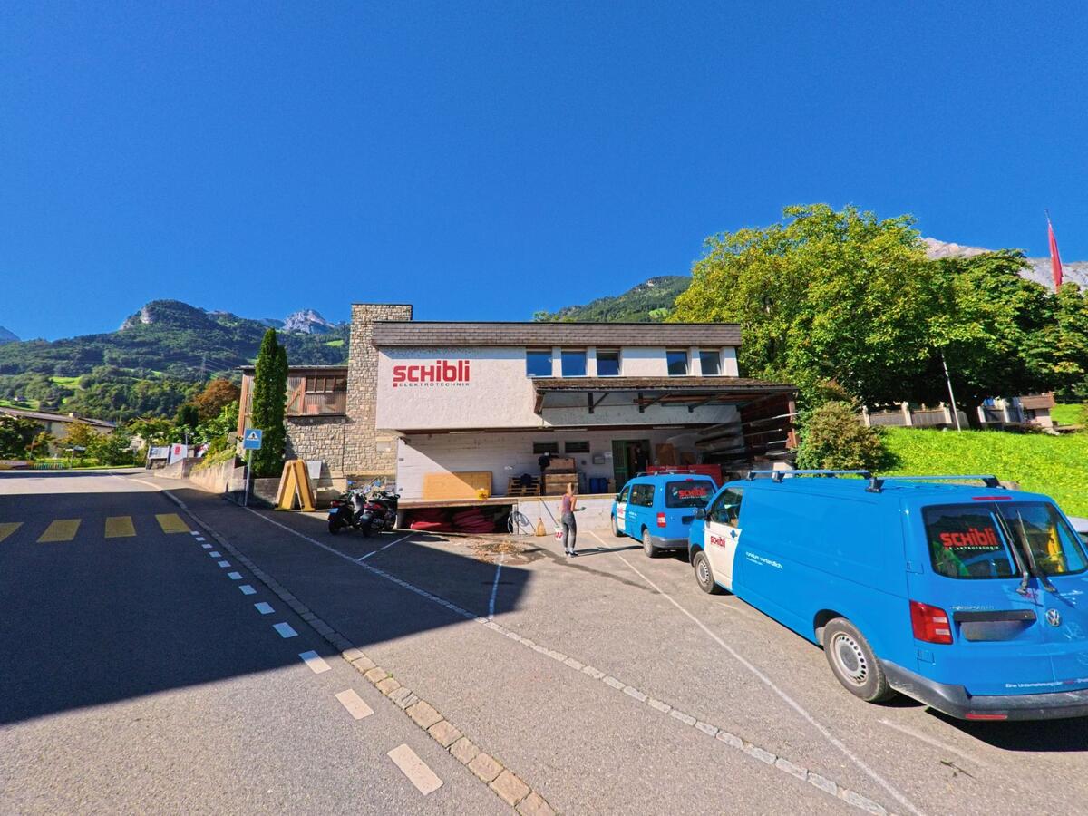 Vor einem Gewerbegebäude der Firma Schibli in Attinghausen (6468, Kanton Uri) stehen mehrere blaue Lieferwagen, im Hintergrund erheben sich bewaldete Berghänge.