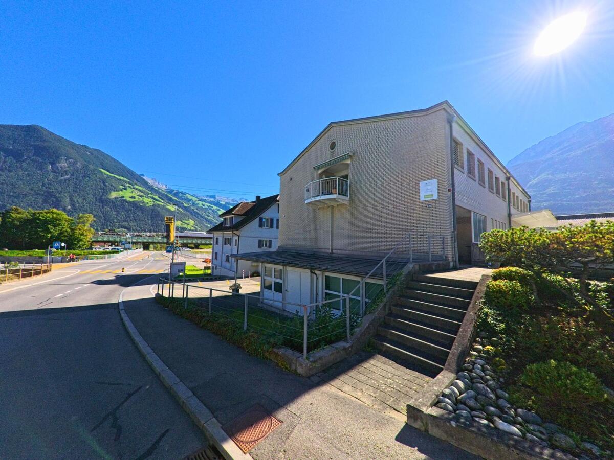 Ein helles Gebaeude mit Treppe und Balkon steht an einer Strasse in Attinghausen (6468, Kanton Uri), waehrend im Hintergrund die Berglandschaft im Sonnenlicht liegt.