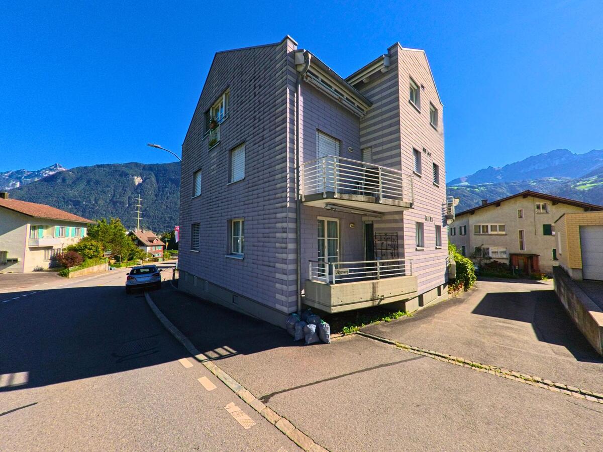 Ein Mehrfamilienhaus mit Balkonen steht an einer Nebenstrasse in Attinghausen (6468, Kanton Uri), umgeben von weiteren Wohnhaeusern und Bergkulisse im Hintergrund.
