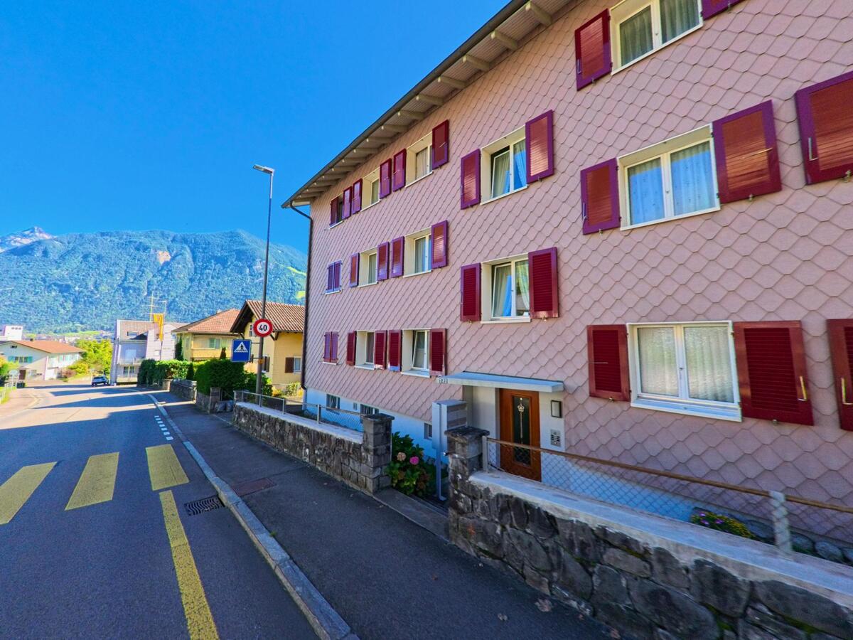 Ein rosa Mehrfamilienhaus mit roten Fensterlaeden steht an einer Strasse in Attinghausen (6468, Kanton Uri), im Hintergrund sind Berge und weitere Wohnhaeuser zu sehen.