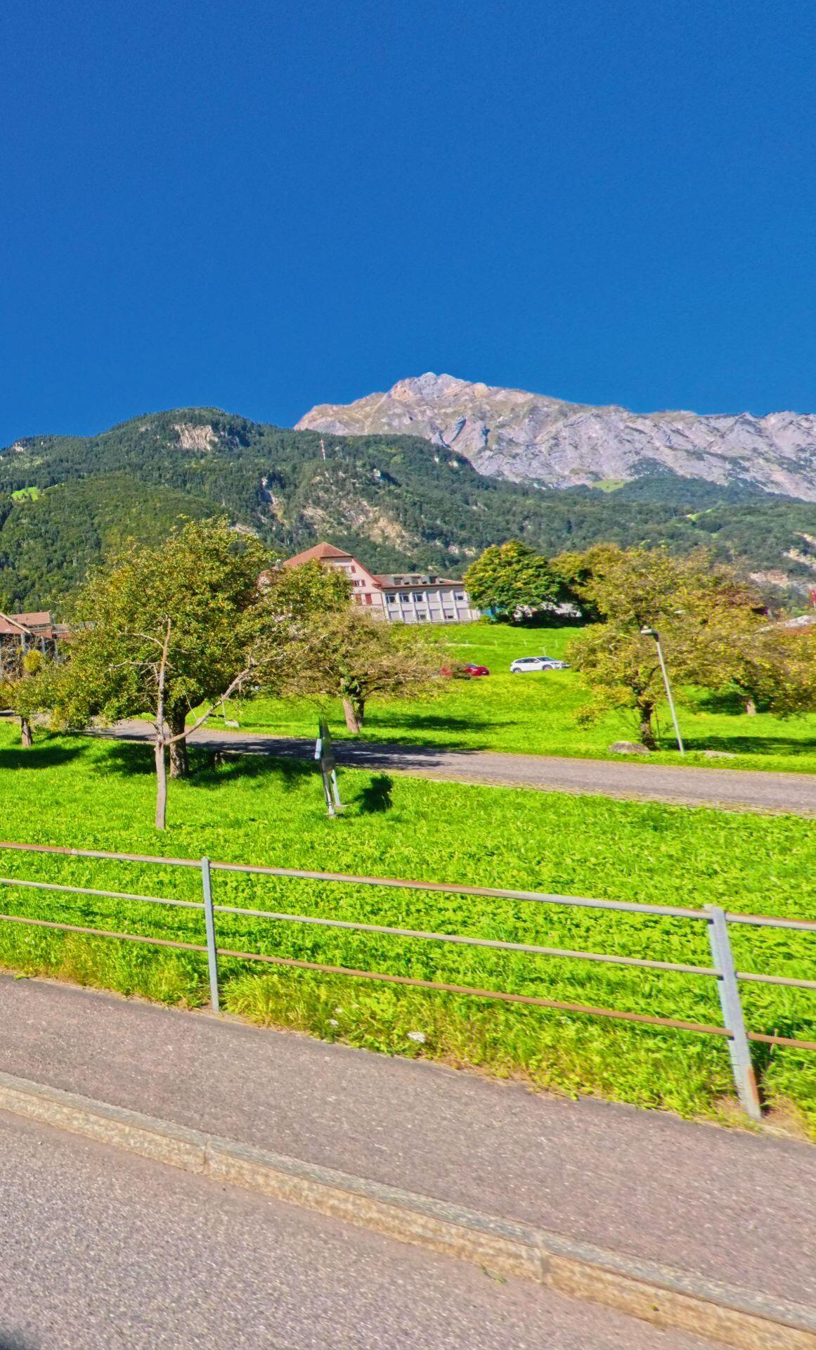 In Attinghausen (6468, Kanton Uri) liegt ein grosses Gebaeude oberhalb einer grünen Wiese mit Obstbaeumen, dahinter ragen die steilen Felswaende der umliegenden Berge empor.