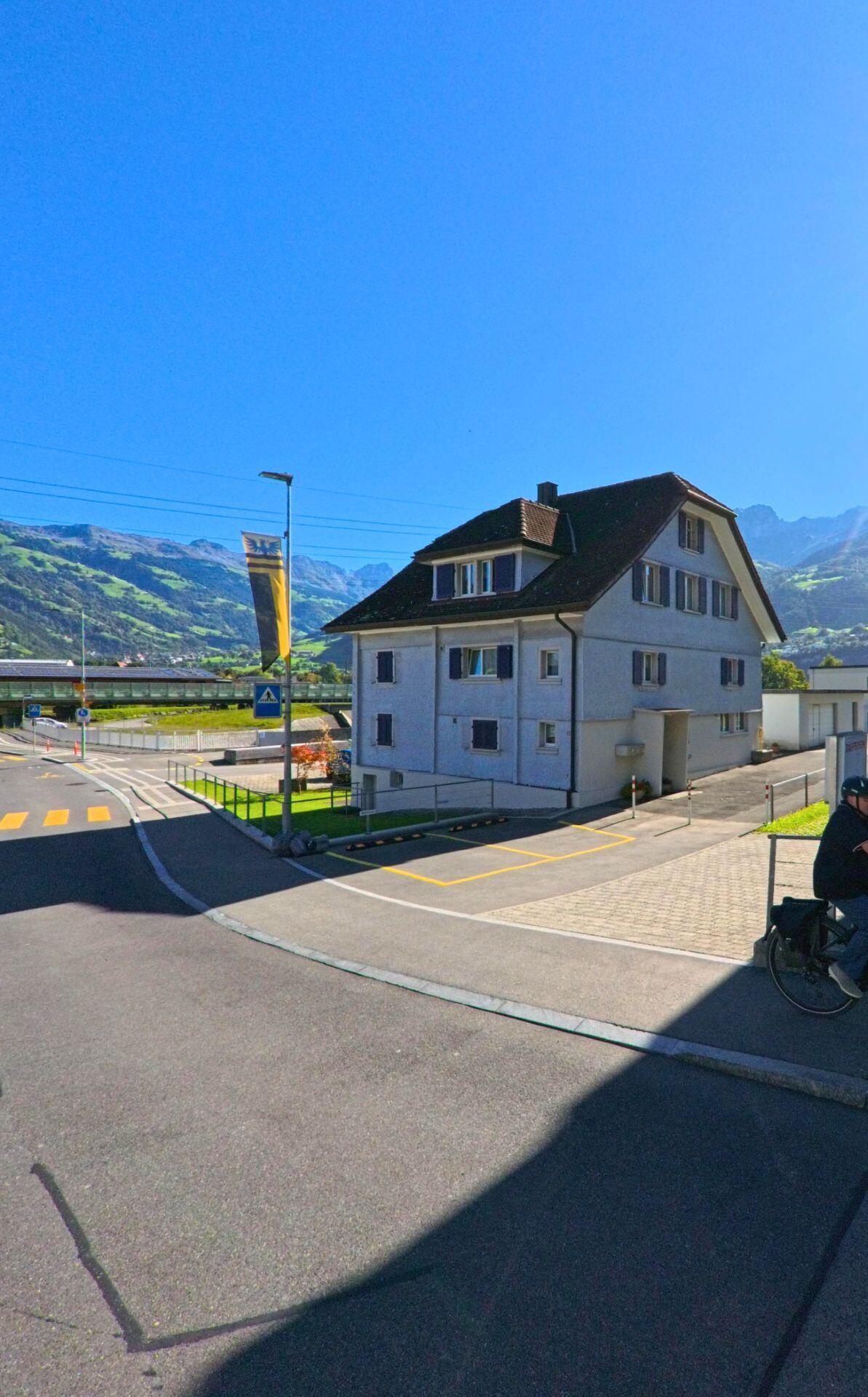 Ein helles Wohnhaus mit Schindeldach steht in Attinghausen (6468, Kanton Uri) an einer ruhigen Strasse mit Blick auf die umliegenden Berge und den Bahnhof im Tal.