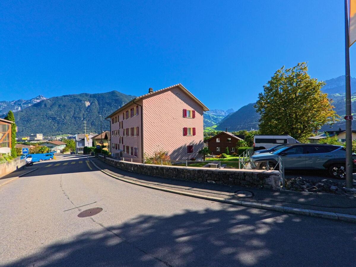 Zwei gelbe Wohnhaeuser mit braunen Fensterlaeden stehen in Attinghausen (6468, Kanton Uri) vor der Kulisse der umliegenden Berge unter strahlend blauem Himmel.
