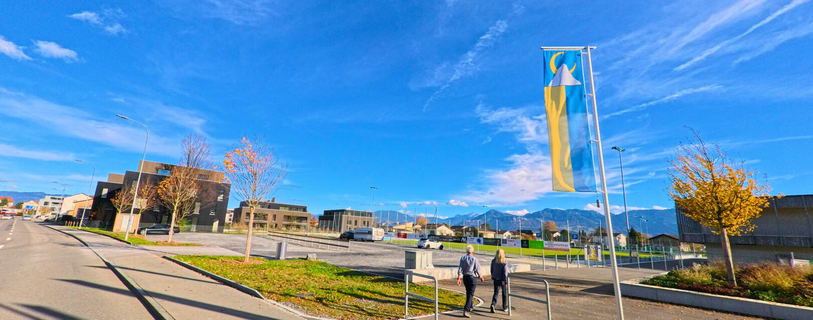 In Au (9434, Kanton St. Gallen) führt eine breite Strasse zwischen modernen Gebäuden vorbei, mit einem klaren Blick auf den Himmel und die Berge im Hintergrund.