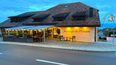 Die Auberge de l’Union in Arzier-Le Muids (1273, Kanton Waadt) zeigt eine gemütlich beleuchtete Fassade mit überdachter Terrasse und rustikalem Dach bei Abendstimmung.