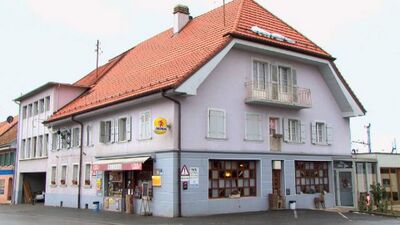 Das Gebäude der Auberge de l'Hôtel de Ville in Semsales (1623, Kanton Freiburg) zeigt eine helle Fassade mit roten Dachziegeln und mehreren Fensterläden entlang der Hauptstrasse.