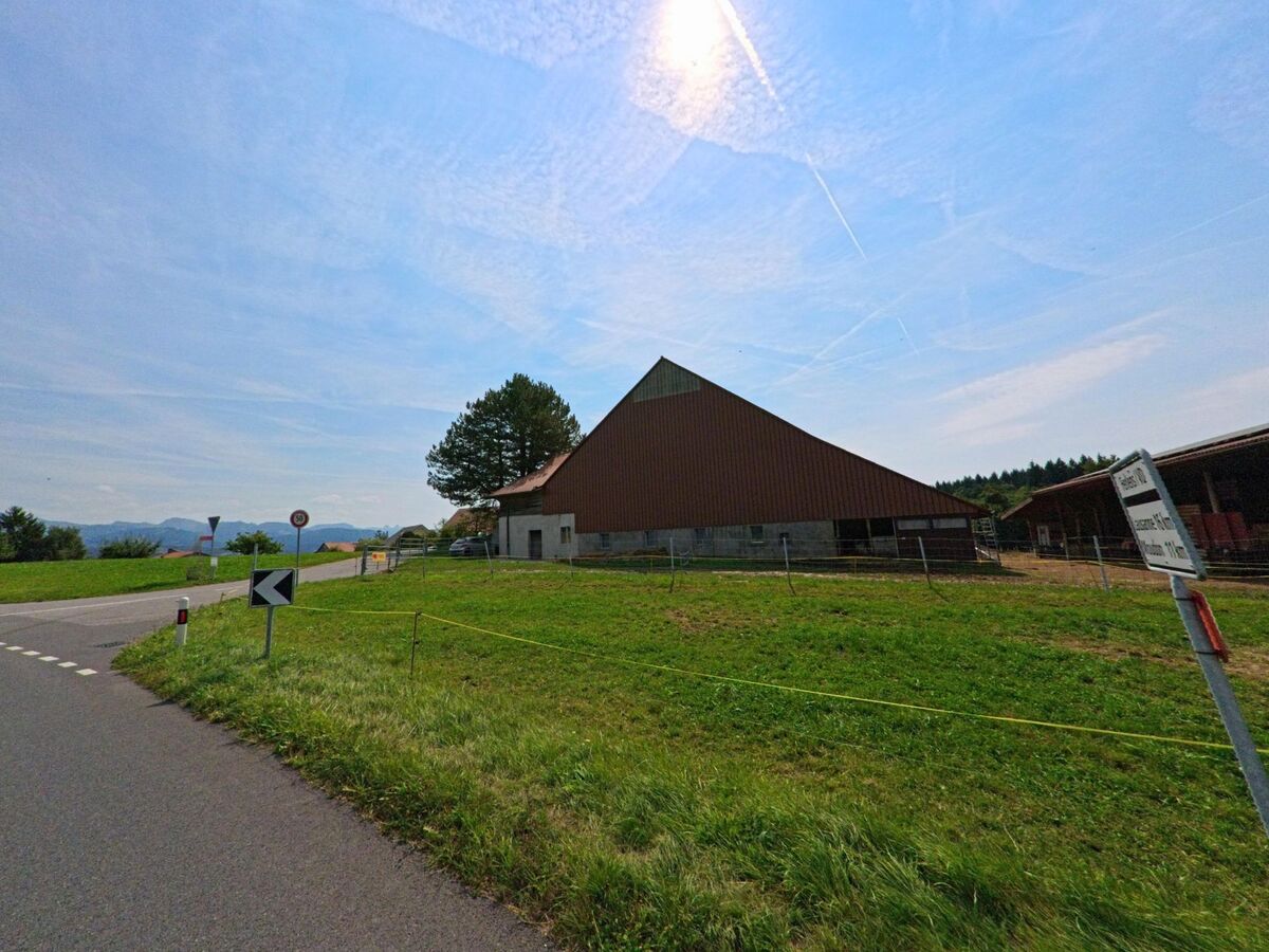 Nahe einer Strassenkurve stehen landwirtschaftliche Gebäude mit umzäunter Wiese in Auboranges (1673, Kanton Freiburg) unter weitem Himmel.