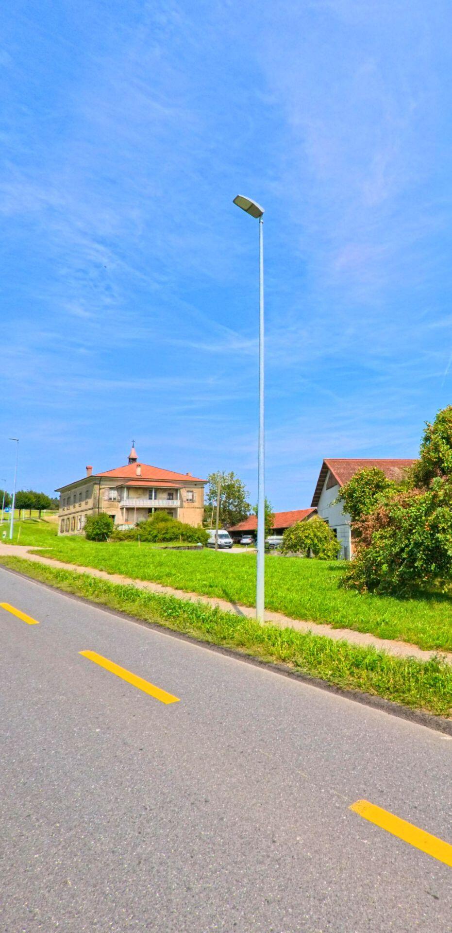 Neben einer ruhigen Strasse mit Gehweg steht ein markantes Gebäude mit Vorplatz in Auboranges (1673, Kanton Freiburg) zwischen Grünflächen und Wohnhäusern.