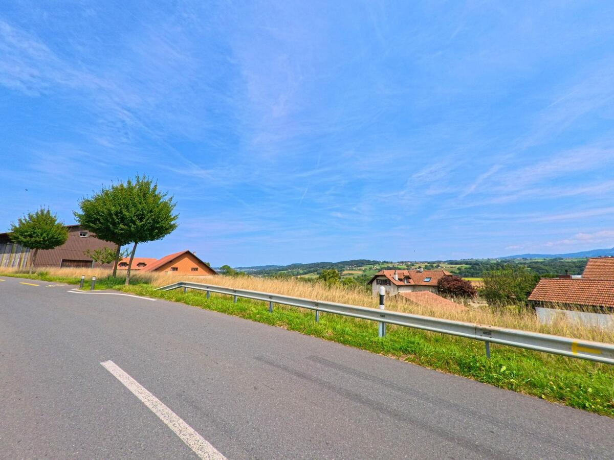 Entlang einer Kurve mit Leitplanke öffnet sich der Blick auf verstreute Wohnhäuser und Hügelland in Auboranges (1673, Kanton Freiburg) unter weitem Himmel.