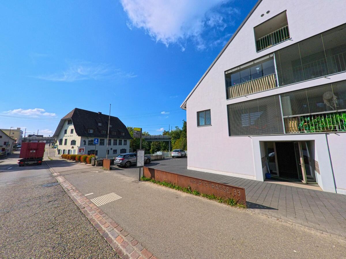 In Augst (4302, Kanton Basel-Landschaft) trifft ein modernes Wohngebaeude mit grossen Fensterfronten auf ein traditionelles Haus gegenueber der Strasse, waehrend ein Lastwagen vorbeifaehrt.