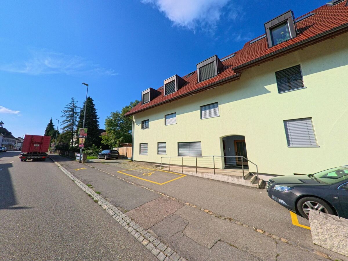 In Augst (4302, Kanton Basel-Landschaft) saeumt ein helles Wohngebaeude mit rotem Dach die ruhige Strasse, waehrend ein Lastwagen und mehrere parkende Fahrzeuge das Bild abrunden.