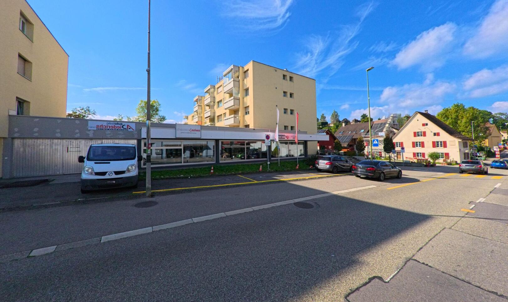 In Augst (4302, Kanton Basel-Landschaft) praegt ein modernes Wohnhaus mit Geschaeftszeile die Strassenszene, waehrend daneben traditionelle Haeuser und parkende Autos das Ortsbild bestimmen.