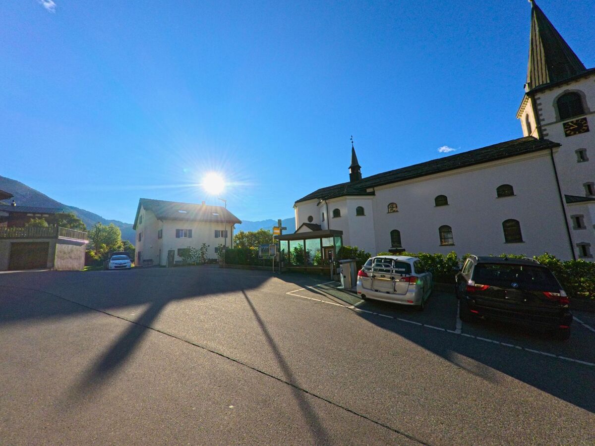 Die Kirche von Ausserberg (3938, Kanton Wallis) wird am Abend von tiefstehender Sonne beschienen, daneben stehen parkende Autos.