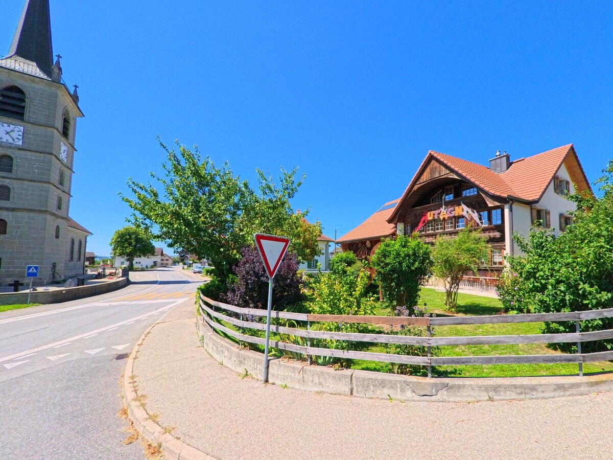 Neben der Kirche von Autigny (1742, Kanton Freiburg) steht ein traditionelles Wohnhaus mit rotem Satteldach und gepflegtem Garten.